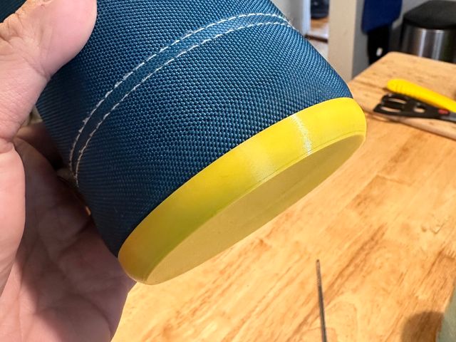 A hand holding the same travel mug, now with a yellow plastic object covering the base. The woven sides are tucked neatly into it and no fraying is visible.