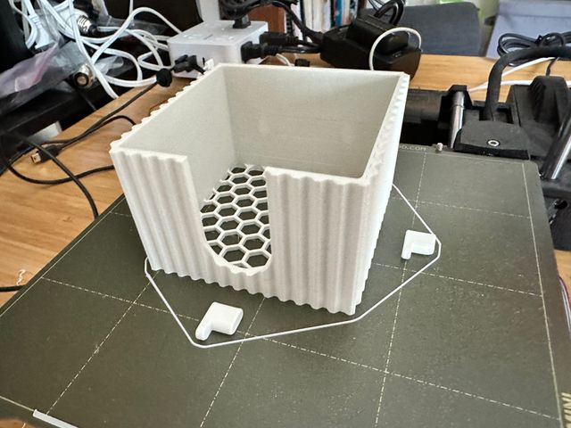 The white sticky note holder on the print bed. It has been printed in three separate parts—two small pegs that look oval feet attached to short legs and the box for holding the notes.