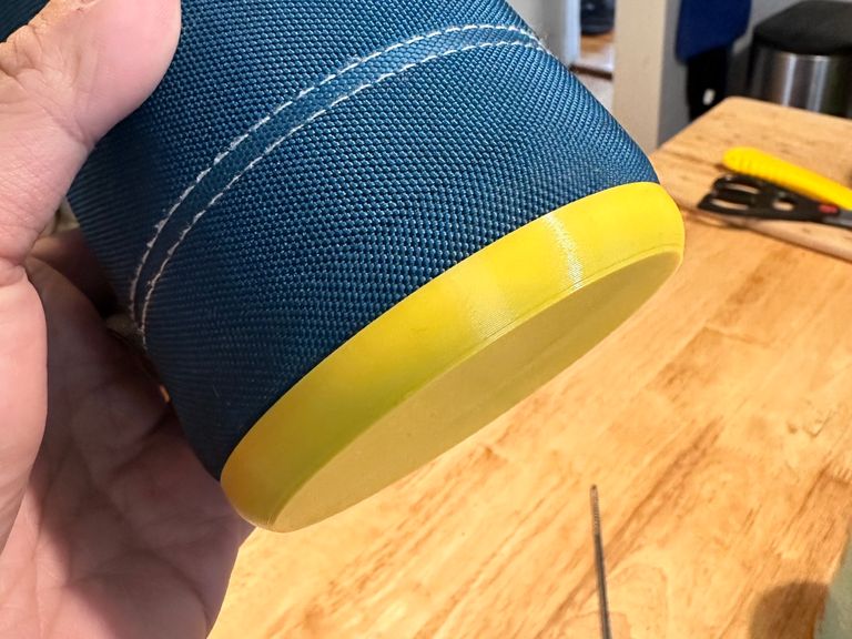 A hand holding the same travel mug, now with a yellow plastic object covering the base. The woven sides are tucked neatly into it and no fraying is visible.