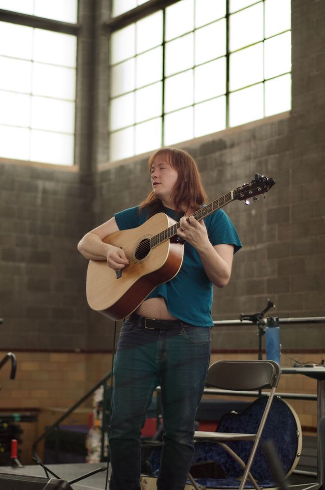 A long haired redheaded guitar player, wearing a cropped blue t shirt, standing and playing an acoustic guitar on stage.