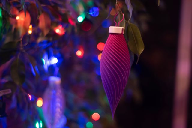 An hanging ornament, shaped like an elongated lemon, featuring a spiral pattern running down its length. It's coloration is a gradient from pink to purple. Behind it, outside of the depth of field, are blurry lights and tree leaves.