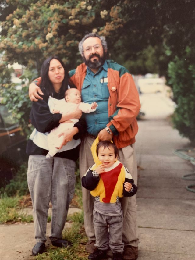 A family of four on a sidewalk with greenery behind them. A man with dark hair and a beard wearing an orange jacket tends to a young toddler in a yellow and red striped outfit. Beside him a woman with dark hair holds a newborn baby.
