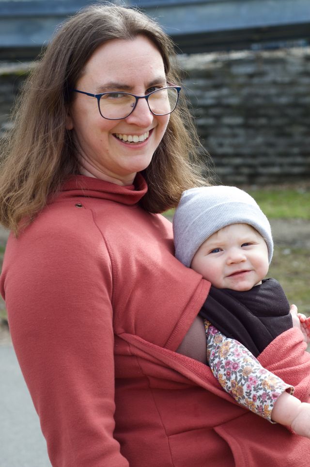 A smiling brown-haired woman with a baby strapped to her chest. The image has a soft quality but individual hairs are crisply rendered even at the edges.