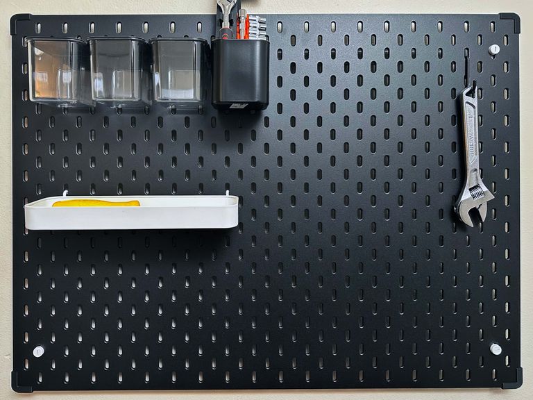 An IKEA Skådis board mounted on a wall. There are boxes and trays mounted on the board and an adjustable wrench hangs from a hook. On each corner of the board is a black bumper.