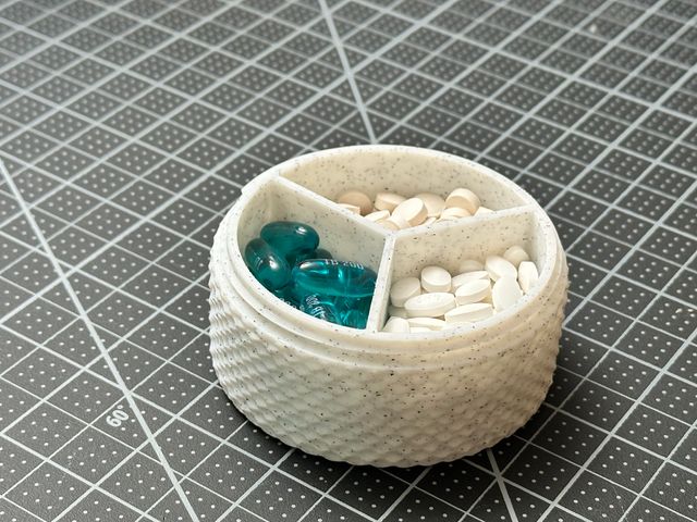 A cylindrical pill case divided into thirds, pie-chart style, with each compartment filled with white or blue pills. The exterior has a knurled texture. The color is white with gray speckles, effecting a marbled look.