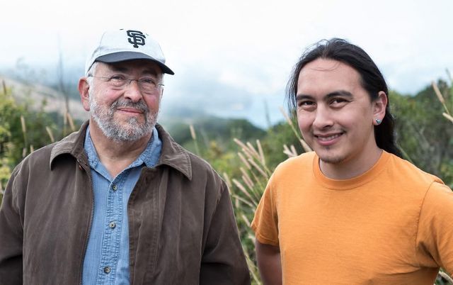 Two men smiling at the camera outdoors. On the left, an older man with a white beard and glasses wearing a brown jacket and an SF Giants baseball cap. On the right, a younger man with long dark hair, a small teal earring, and an orange t-shirt. Misty hills and tall grasses are visible behind them.