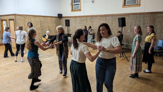 A room with contra dancers holding two hands with their partner, in motion. In the background I'm calling with a band.