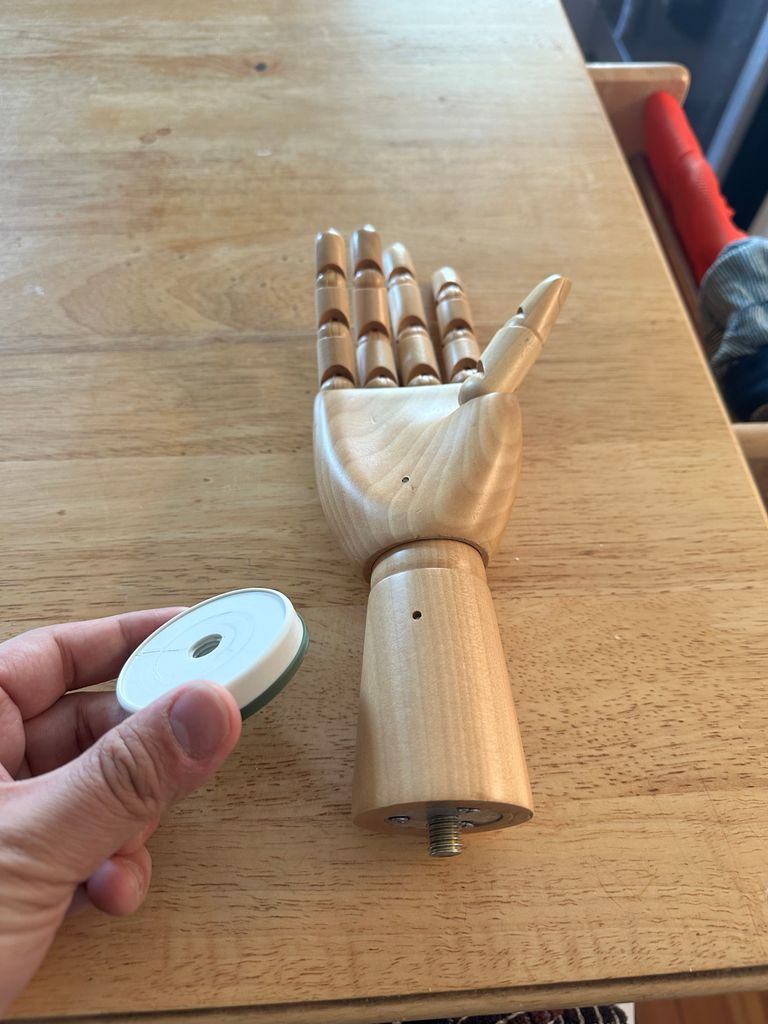 A wooden articulated hand (like for drawing) laying on a counter. There is a metal threaded rod extending from the flat base of it, below the wrist. On the left side of the hand is a (real) hand holding a green and white plastic cylinder with a hole in the middle.