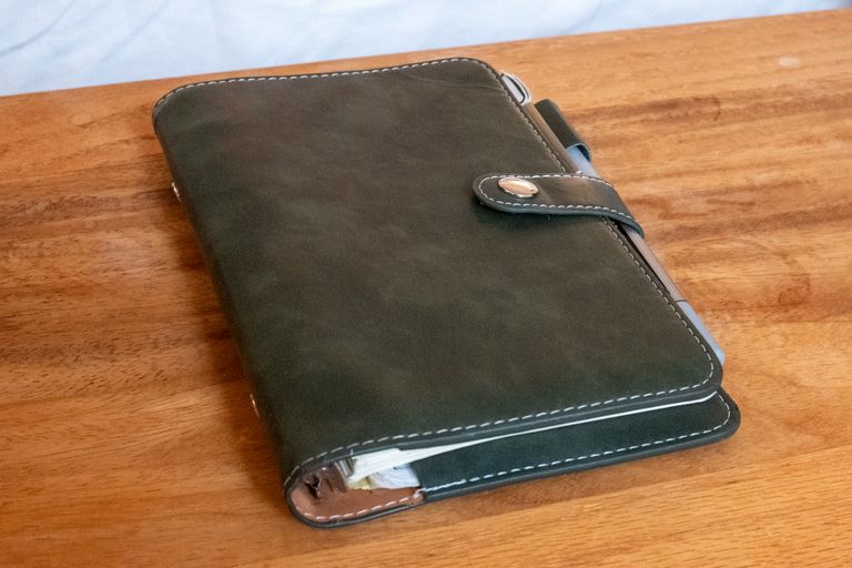 The closed journal on a wooden table: a dark green faux-leather cover with contrast stitching, a snap strap, and a pen in the loop along its edge
